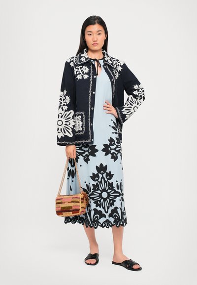 Marineblazer mit floralem Muster und weißen Akzenten, kombiniert mit einem hellblauen Kleid mit schwarzen Designs. Braune geflochtene Tasche und schwarze Sandalen.