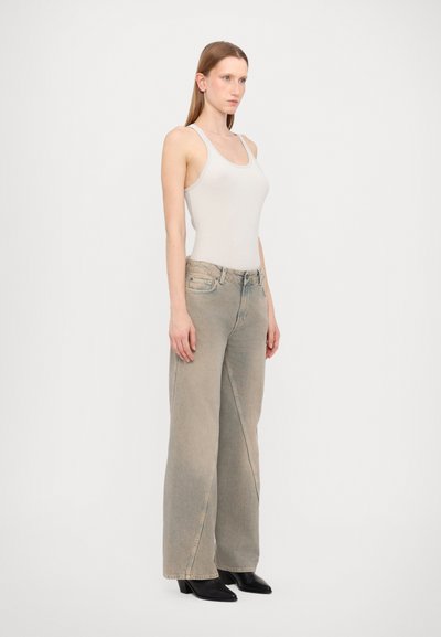 Hellgraues geripptes Tanktop kombiniert mit weit geschnittenen, verblichenen Jeans. Schwarze Stiefeletten mit Absatz vervollständigen das Outfit. Das Model präsentiert ein Profil.