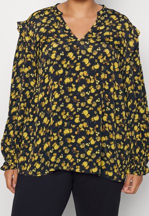 Løs sort bluse med gult blomsterprint, flæser på skuldrene og rynkede manchetter, båret med sorte bukser.