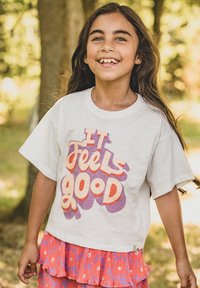 Wit T-shirt met korte mouwen en kleurrijke "IT FEELS GOOD"-grafiek in paars en oranje, gecombineerd met een gelaagde roze rok met bloemenpatronen.