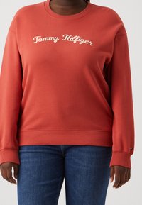 Personne portant un sweatshirt couleur orange brûlé de Tommy Hilfiger avec un texte de logo blanc et un jean bleu, les mains détendues le long du corps.