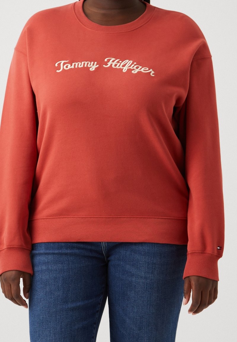 Personne portant un sweatshirt couleur orange brûlé de Tommy Hilfiger avec un texte de logo blanc et un jean bleu, les mains détendues le long du corps.