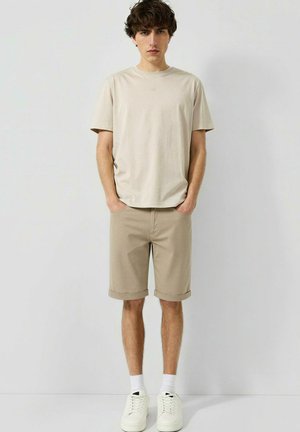 Jeune homme debout les mains dans les poches, portant un t-shirt beige, un short assorti, des chaussettes blanches et des baskets blanches sur un fond uni.