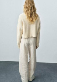 Maglione a collo alto di colore crema abbinato a pantaloni larghi in lino beige chiaro, che mostrano un fit rilassato e un tessuto testurizzato.