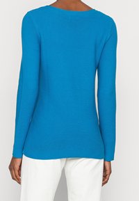 Haut bleu à manches longues en tissu côtelé texturé, col arrondi et manches ajustées. Associé à un pantalon de couleur claire.