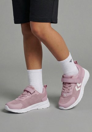 Dziecko w białych skarpetkach i różowych sportowych butach z białymi podeszwami stoi na szarej powierzchni na tle jednolitego, szarego tła.