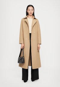 Beige doppelt knöpfter Mantel mit einem Reverskragen, langen Ärmeln und lockerer Passform, kombiniert mit schwarzen, weit geschnittenen Hosen und einer kleinen schwarzen Handtasche.