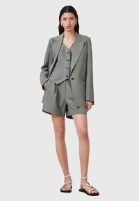 Blazer grigio chiaro su misura e gilet coordinato con bottoni, abbinati a pantaloni corti a vestibilità ampia e sandali con cinturini. Tessuto di trama liscia.