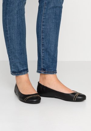 Person iført mørkeblå skinny jeans og sorte læderballerinaer med gyldt Tommy Hilfiger-logo på tæerne, stående på lys gulv.