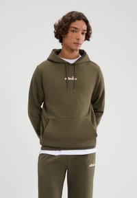 Oliwkowa bluza z kapturem z kieszenią kangurzą, z białym logo na piersi, kapturem na sznurki i ściągaczami na rękawach. Połączona z pasującymi spodniami dresowymi.