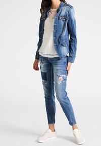 Jeansjacka med nitar, över en vit spetsblus. Tärda jeans med blommönster. Ljusfärgade sneakers fulländar outfiten.