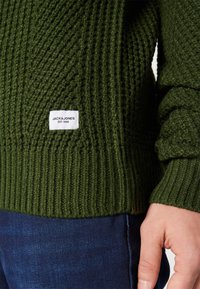 Dark green textured knit sweater with ribbed cuffs, featuring a small white label with branding near the hem. Paired with dark blue jeans.