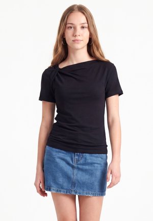 Jeune femme aux longs cheveux portant des boucles d'oreilles en anneau, un haut noir à manches courtes et une jupe en jean bleue, debout devant un fond blanc.