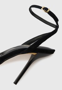 Črne semiš sandale z visoko peto, s tankim trakcem okoli gležnja in z zlatim zaklepom. Imajo koničast nos in elegantno stiletto peto.