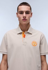 Beige polo skjorte i struktureret stof med en to-knaps knapstolpe, orange kraveaccent og rundt logo på venstre bryst.