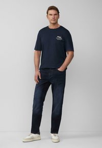 Marineblauwe t-shirt met logo op de voorkant, gecombineerd met donkerblauwe spijkerbroek en witte sneakers. De stof lijkt zacht, met een casual pasvorm en strakke lijnen.