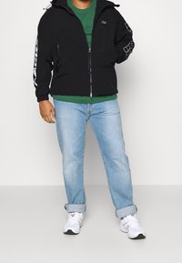 Svart zip-up jacka med huva, med vitt logotyp och geometriskt mönster på ärmarna. Matchad med en grön skjorta och ljusblå jeans.