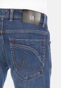 Jeans de mezclilla azul con costuras naranjas, que cuentan con un parche de cuero negro etiquetado como "LTB" y dos bolsillos traseros con curvas distintivas.