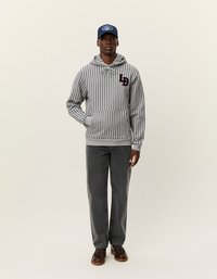 Pelēka svītraina kapuce džemperis ar sarkanu logo uz krūškurvja, savienots ar pelēkiem džinsiem un brūnām kurpēm, zem tumši zilā cepures.