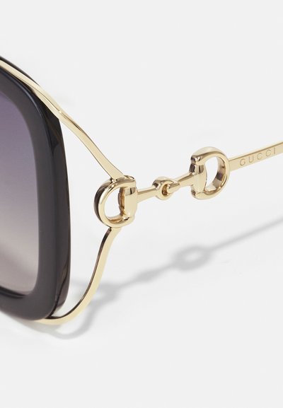Lunettes de soleil Gucci avec des verres dégradés noirs, des montures en métal doré et des accents en matériel circulaire. Texture lisse et design moderne.