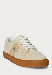 Beige suede sneaker with white laces, tan accents, and textured rubber outsole. Features a small logo on the side and a rounded toe design.