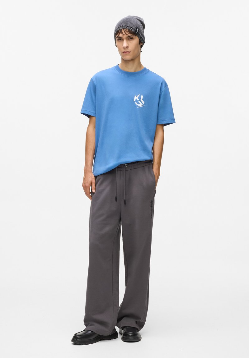 Zila kokvilnas T-kreklā ar priekšējo logo, pelēkas sporta bikses ar sānu kabatām un melnu kordu apavu. Modelis valkā pelēku cepuri.