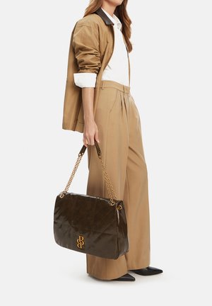 Kobieta ubrana w beżowe spodnie i marynarkę z podwiniętymi rękawami, trzymająca dużą ciemnobrązową pikowaną torebkę na złotych łańcuszkach.