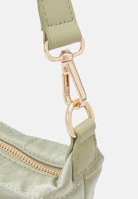 Gros plan sur le coin d'un sac matelassé vert clair avec une fermeture éclair dorée, un fermoir en or et une sangle verte assortie attachée par un anneau doré.