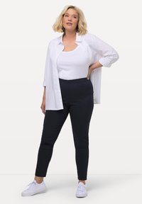 Bílá košile na knoflíky přes bílý tílko, v kombinaci s tmavě modrými kalhotami a bílými teniskami. Tento outfit zdůrazňuje neformální a pohodlný styl.