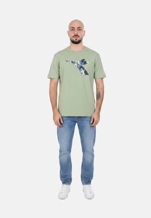 Uomo con t-shirt verde chiaro con logo mimetico, jeans blu, sneakers bianche e un tatuaggio sull'avambraccio sinistro, in piedi davanti a uno sfondo uniforme.