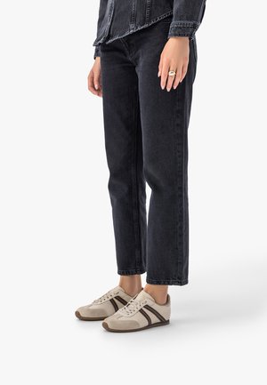 Osoba nosząca ciemne, proste jeansy i beżowe trampki z brązowymi paskami, stojąca na jednolitym białym tle.