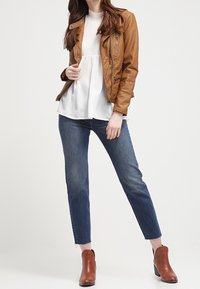 Kobieta ubrana w brązową skórzaną kurtkę, białą bluzkę, niebieskie dżinsy i brązowe botki za kostkę, stojąca na tle jednolitego tła.