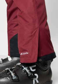 Bordeaux ski pants met een gladde structuur, voorzien van zijzakken en een zwarte accentboord. Gecombineerd met zwarte skischoenen en metalen gespen.