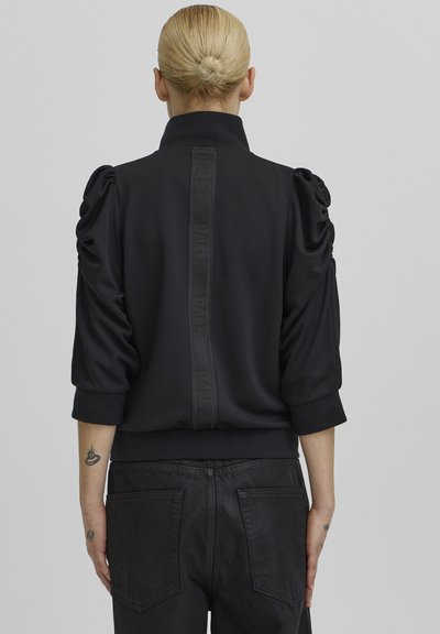 Crni pulover s napuhanima rukavima i stojećim ovratnikom; sadrži vertikalni teksturirani panel s logotipom na leđima. Glatki materijal.