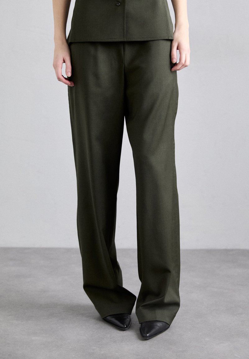 Calças largas de cor verde-oliva em tecido suave, com uma cintura alta e drapeados subtis, combinadas com saltos pretos pontiagudos.
