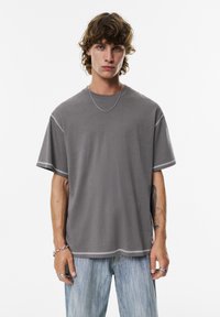 Grå kortærmet t-shirt med en afslappet pasform, prydet med kontrasterende syninger, rund hals og blødt stof. Kombineret med lyseblå jeans.