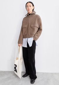 Brun kort jacka med hög krage, två framfickor, bärs över en ljusblå skjorta, kombinerad med svarta vida byxor och platta skor.