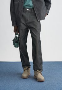 Jeans rectos de mezclilla oscura con costuras en contraste, combinados con botas beige con cordones y una camisa azul verdoso, sostenidos con un bolso verde.