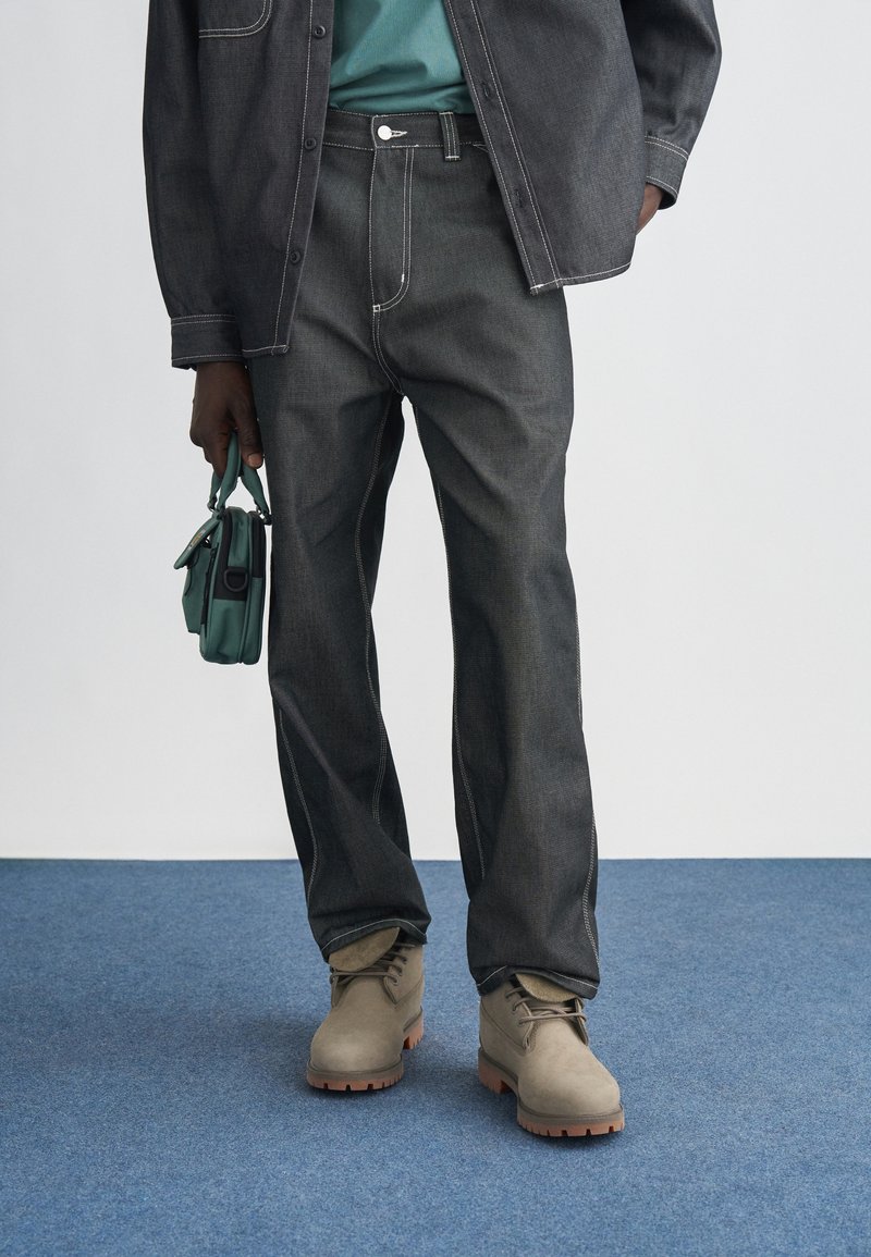 Jeans rectos de mezclilla oscura con costuras en contraste, combinados con botas beige con cordones y una camisa azul verdoso, sostenidos con un bolso verde.