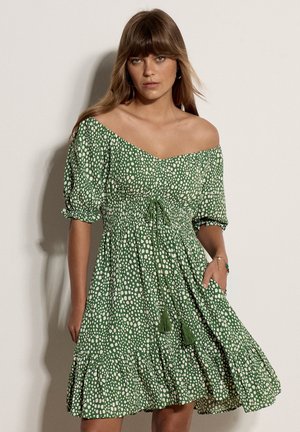 Kvinde iført grøn off-shoulder kjole med hvide prikker, pufærmer og en flæsekant, der står mod en ensartet lys baggrund.