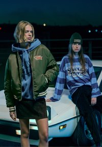 Olivová bombarďák s vyšívaným logom, modrá mikina pod ním, čierna semišová sukňa; druhý model v pruhovanej fialovej a modrej mikine s čiapkou.