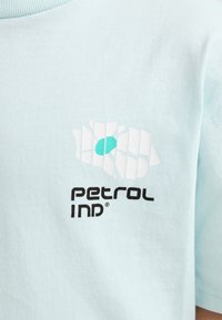 Jasnoniebieska bawełniana koszulka z białym nadrukiem kwiatowego wzoru i turkusowym detalem, z napisem "PETROL IND" w pogrubionych czarnych literach.