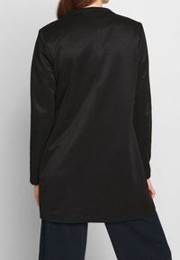 Veste noire à manches longues avec une texture lisse, un design sur mesure et une fente unique à l'arrière. Comprend un col cranté et un style minimaliste.
