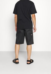 T-shirt in cotone nero e pantaloni corti neri larghi con un patch logo; indossato con sandali intrecciati neri. Sfondo neutro con illuminazione soffusa.