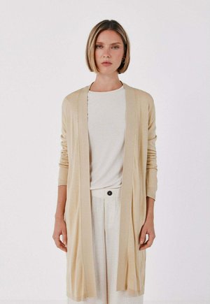 Donna con capelli biondi corti indossa un cardigan lungo beige sopra una maglietta bianca e pantaloni chiari su uno sfondo bianco uniforme.