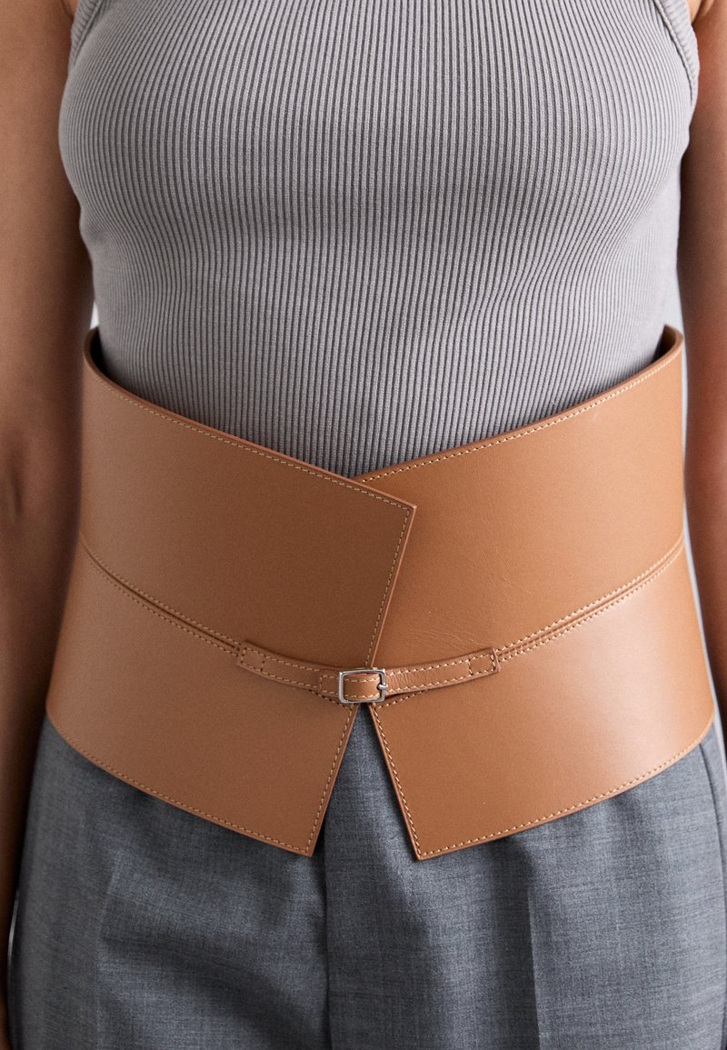 Ceinture en cuir marron, large et structurée, présente un motif croisé et une boucle réglable pour la fermeture. Texture lisse avec des coutures soignées.