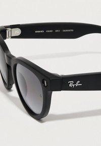Gafas de sol negras con lentes tintados en gris. Presentan un diseño rectangular simple, textura lisa y herrajes plateados. Logo de Ray-Ban en la montura.