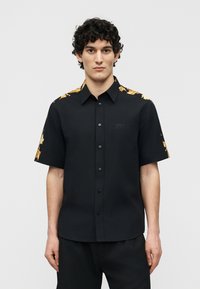 Άνδρας με σγουρά μαλλιά, φοράει μαύρο πουκάμισο με κοντά μανίκια και κουμπιά, με χρυσό μπαρόκ σχέδιο στους ώμους και τα μανίκια.