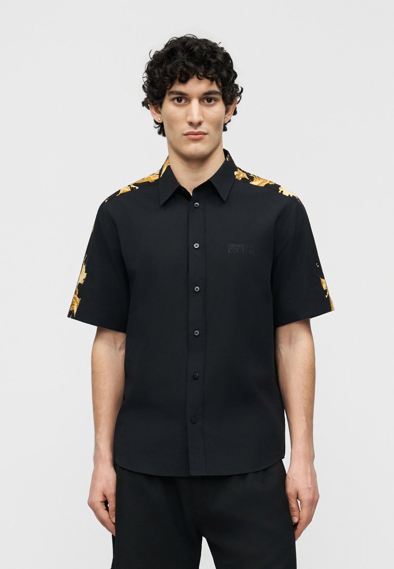 Άνδρας με σγουρά μαλλιά, φοράει μαύρο πουκάμισο με κοντά μανίκια και κουμπιά, με χρυσό μπαρόκ σχέδιο στους ώμους και τα μανίκια.
