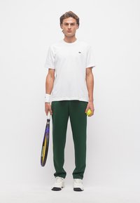 Νέος άνδρας με λευκό μπλουζάκι και πράσινο παντελόνι, κρατώντας ρακέτα τένις και μπάλα, στέκεται μπροστά σε απλό λευκό φόντο.